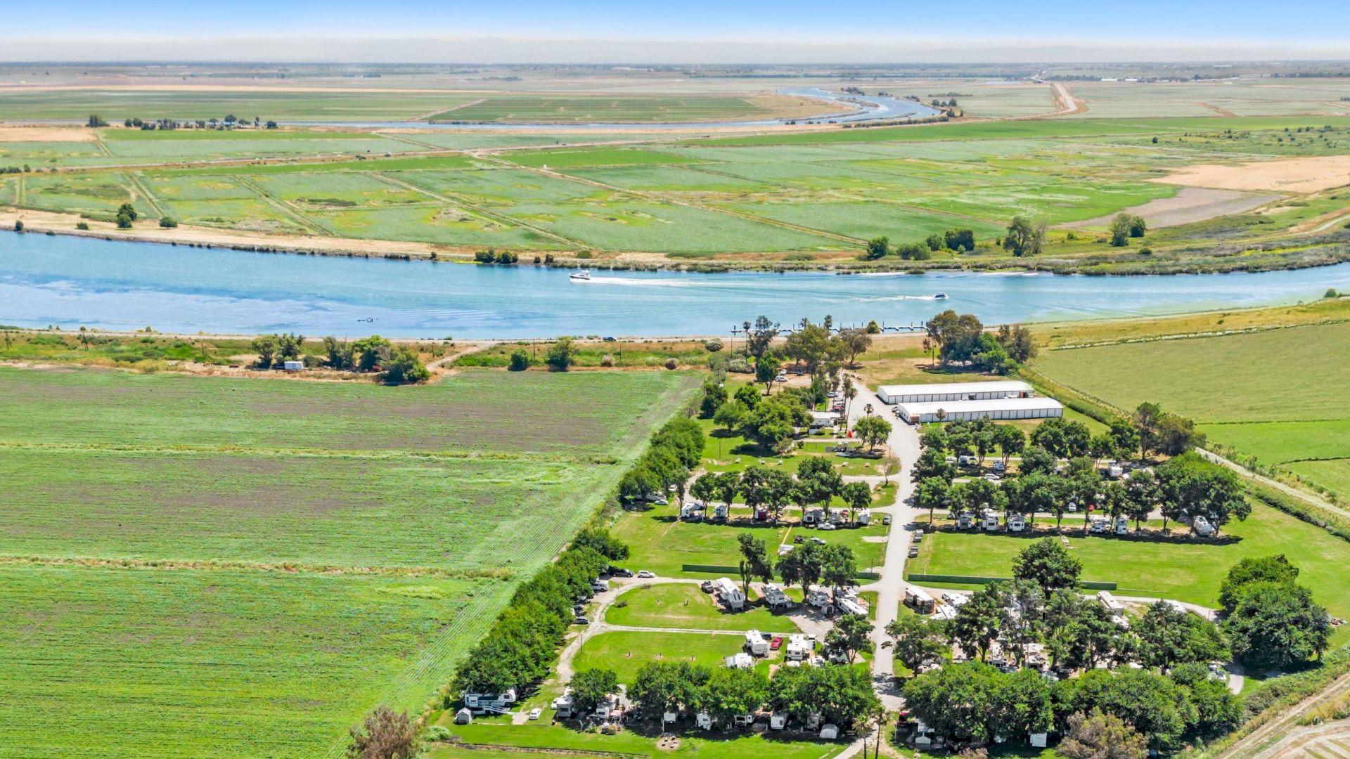Aerial view of a riverside farm with green fields, boats, a small harbor, and a cluster of buildings and RVs.
