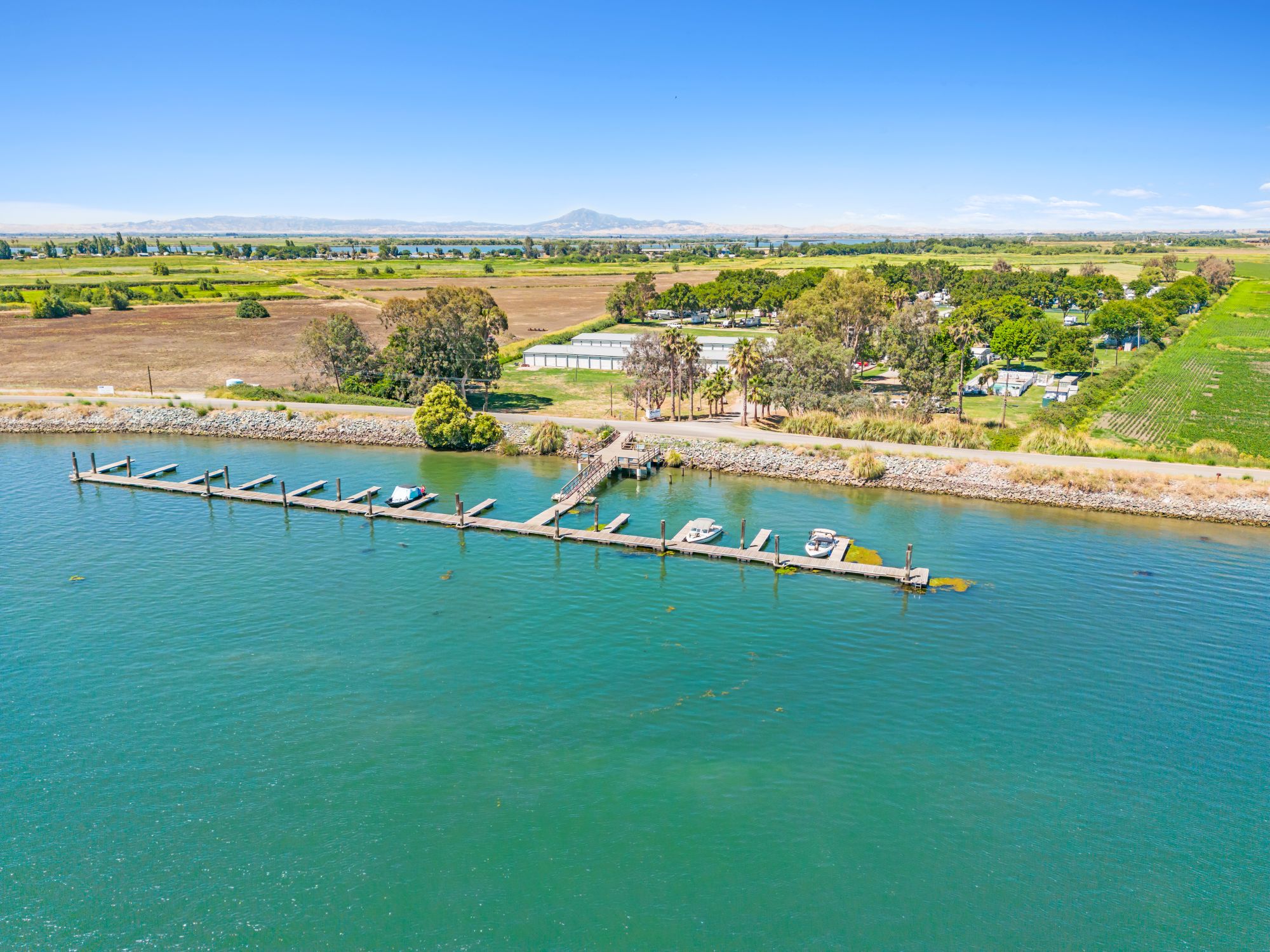 Aerial view of a river with a wooden dock and boats, beside lush fields and a large building under a clear blue sky.