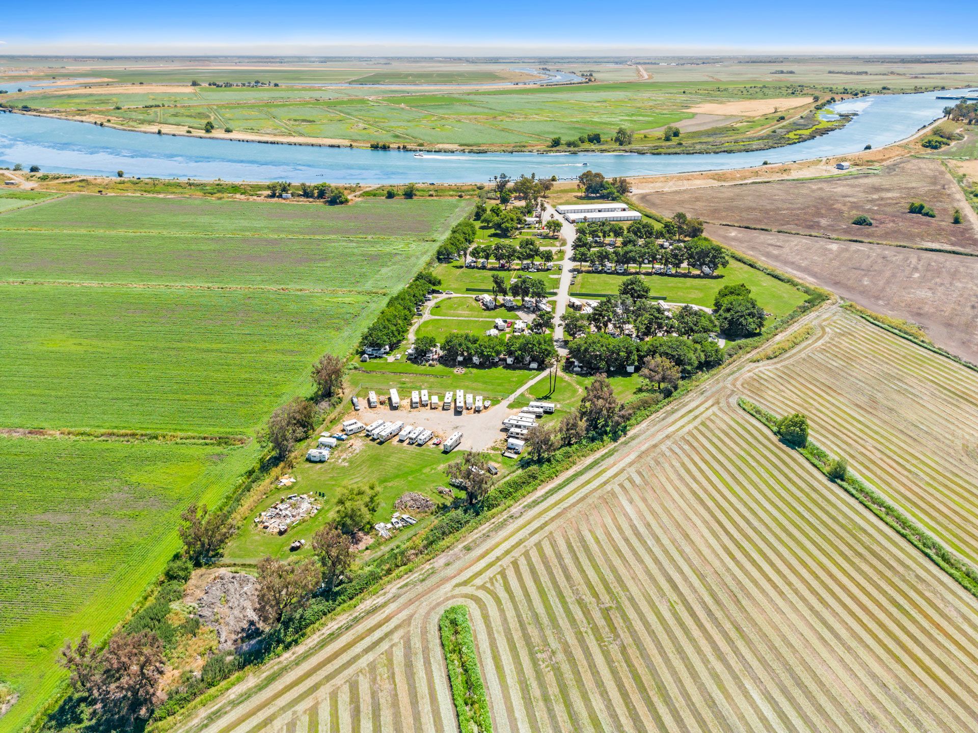 Aerial view of a riverside rural campground and farm fields with caravans, trees, and boats along the water.