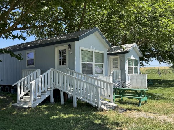 A small white cabin with a railing, set in a grassy area with trees and a picnic table nearby.