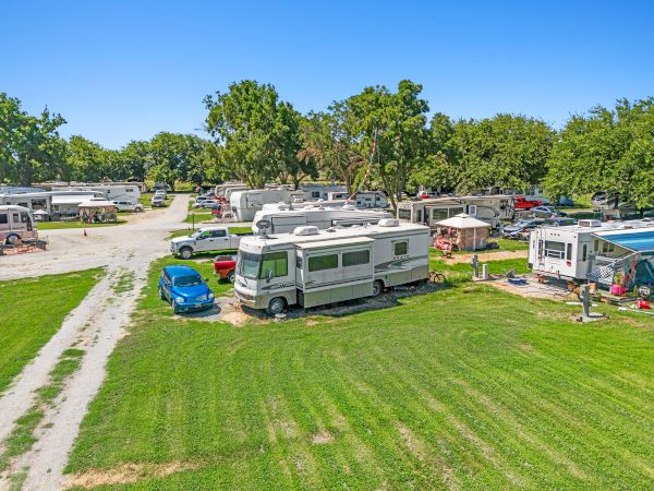 The image shows a campground with several RVs and a few cars on a grassy area with trees in the background under a clear blue sky.