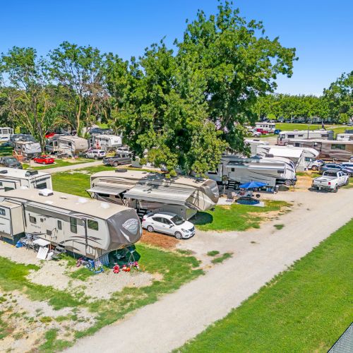 The image shows a campground with multiple RVs and trailers parked among trees and grassy areas under a clear blue sky.