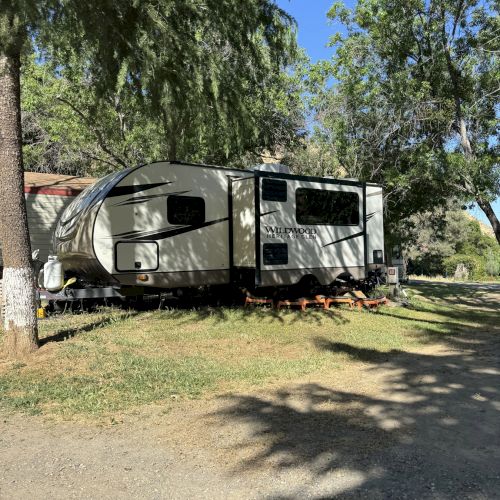 The image shows a travel trailer parked on grass, surrounded by trees, under a clear blue sky.