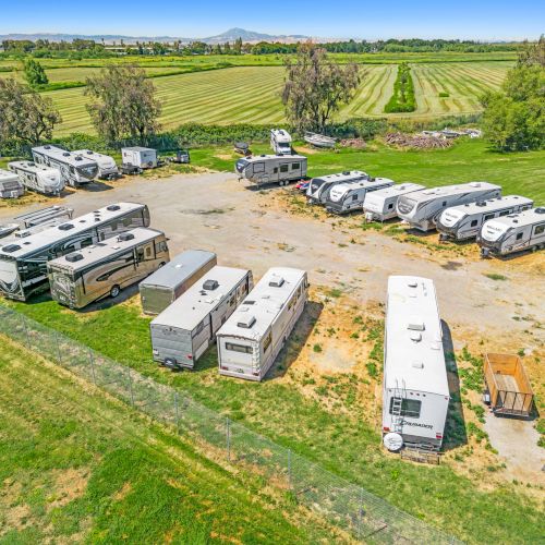 This image shows a lot with several parked RVs surrounded by fields and trees, suggesting a rural or countryside setting.