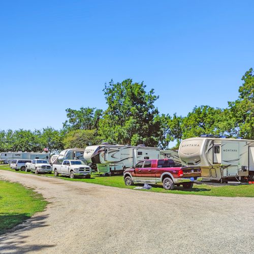 The image shows a campground with multiple RVs parked along a gravel road, surrounded by trees and a clear blue sky.
