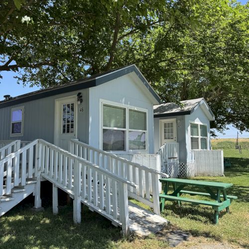A small, light blue house with a white railing and a green picnic table outside, surrounded by trees and grass under a clear sky.
