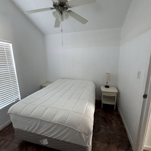 A bedroom with a ceiling fan, bed with white bedding, two side tables, and a lamp, in a room with white walls and blinds on the window.