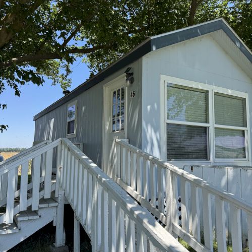 A small, light-colored house with a sloped white railing, front door, and windows is shaded by a tree.