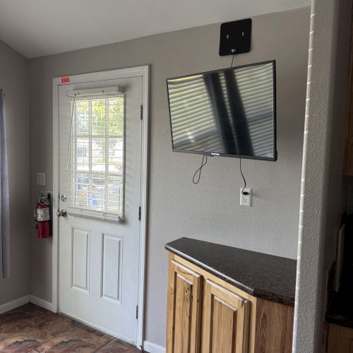A small room corner with a wall-mounted TV, white door, fire extinguisher, and a wooden cabinet on a tiled floor.