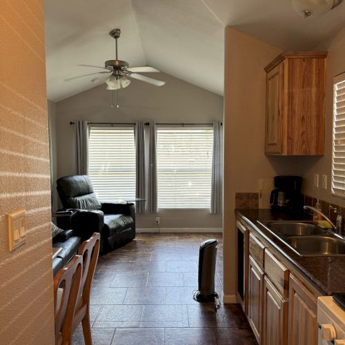 A cozy living space with a kitchen, seating area, ceiling fan, and large windows with blinds, giving a warm and inviting atmosphere.