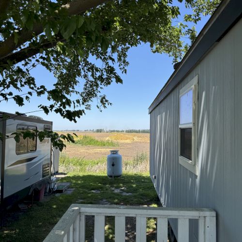 A mobile home and RV are in a grassy area with a tree and a view of a field, under a clear blue sky.