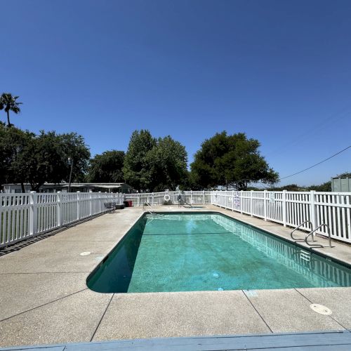 A rectangular swimming pool surrounded by a white fence under a clear blue sky, with trees in the background.