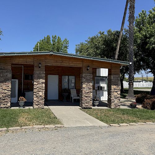 A small, single-story building with a stone facade, flanked by trees and a palm, sits on a grassy area with a sidewalk in front.