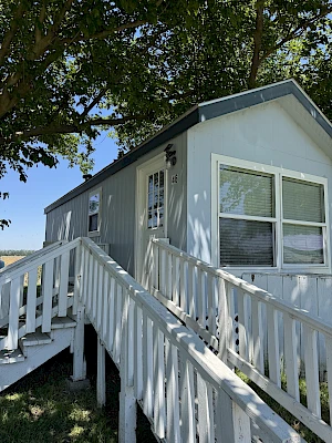 The image shows a small gray house with a white ramp leading up to the door, set under trees and surrounded by a grassy area.