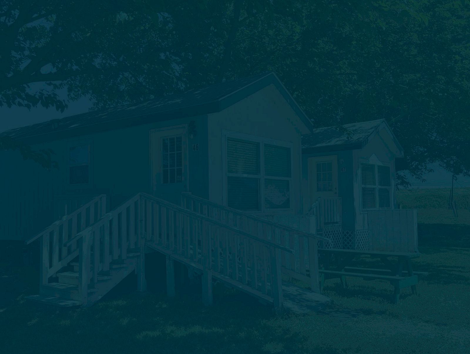 The image shows a small house with a porch and steps leading up to it, surrounded by trees and grass in a shaded setting.