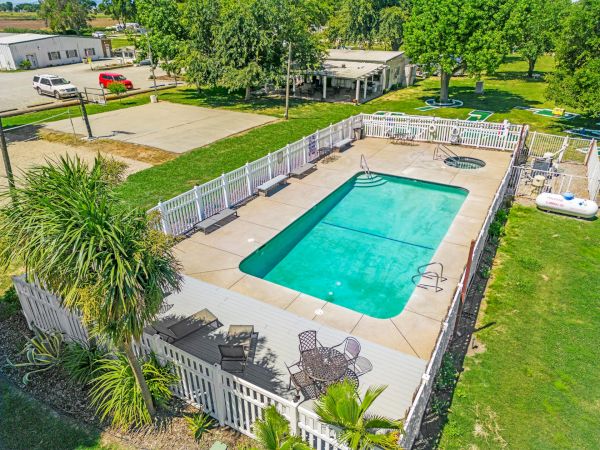The image shows a fenced outdoor swimming pool with surrounding patio chairs and tables, set in a grassy area with trees and nearby buildings.