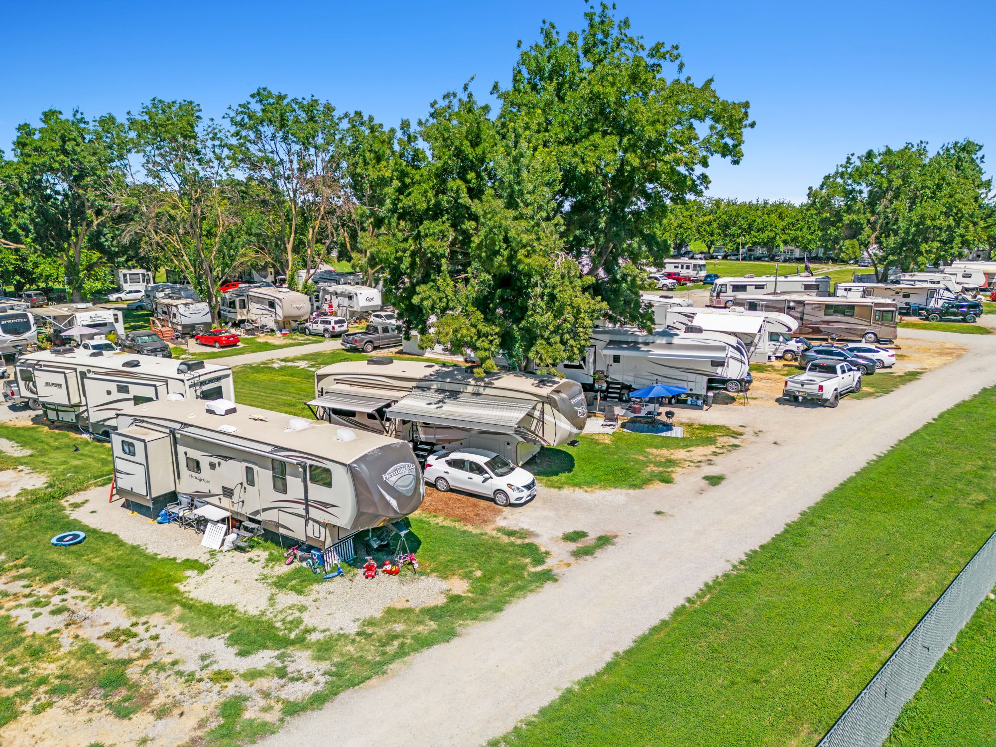 Aerial view of a campground with numerous RVs parked along dirt roads, green grass, and tall trees under a clear blue sky.