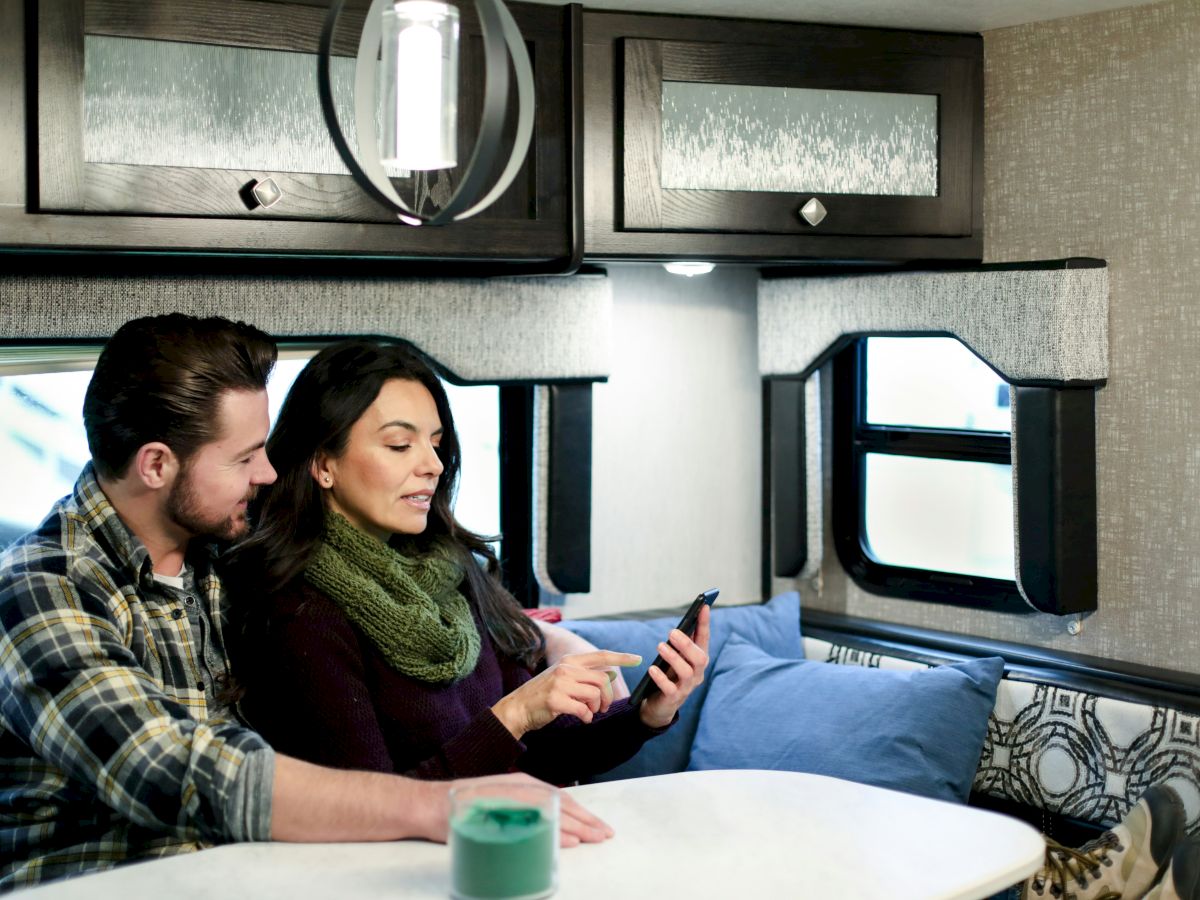 Couple inside a cozy camper, man and woman sit at a round table looking at a tablet; bright windows, cushions, and rustic decor surround them.