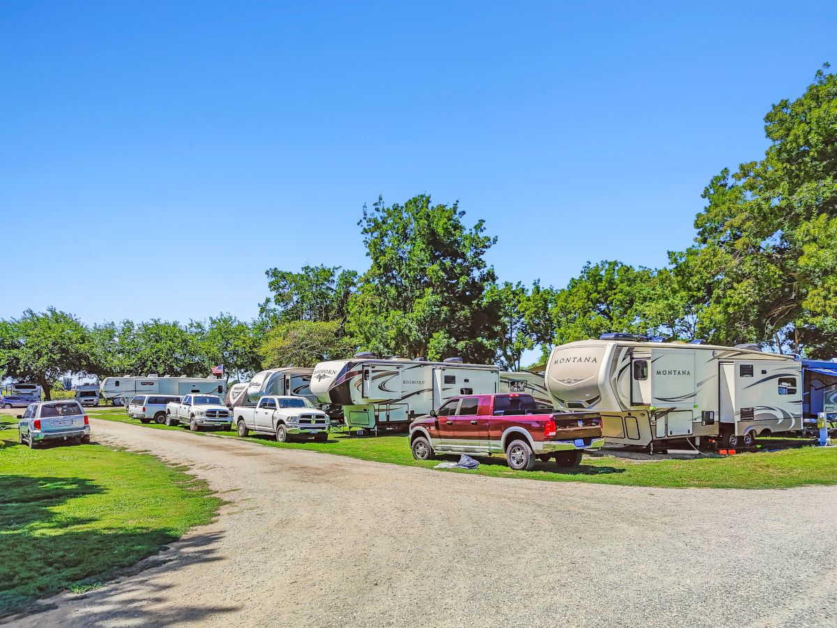 A campground filled with a row of RVs and trailers parked along a gravel road under a bright blue sky, with green trees in the background.