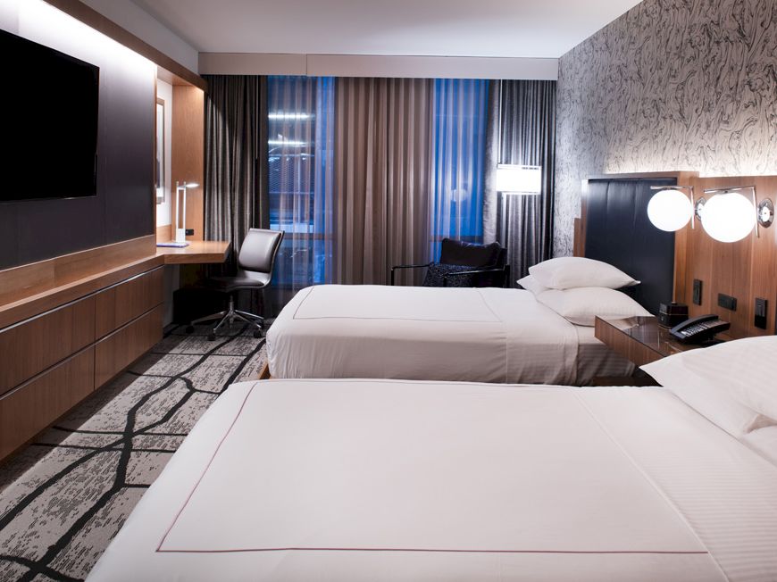 A modern hotel room with two beds, a wall-mounted TV, lamps, and a desk by large window curtains.