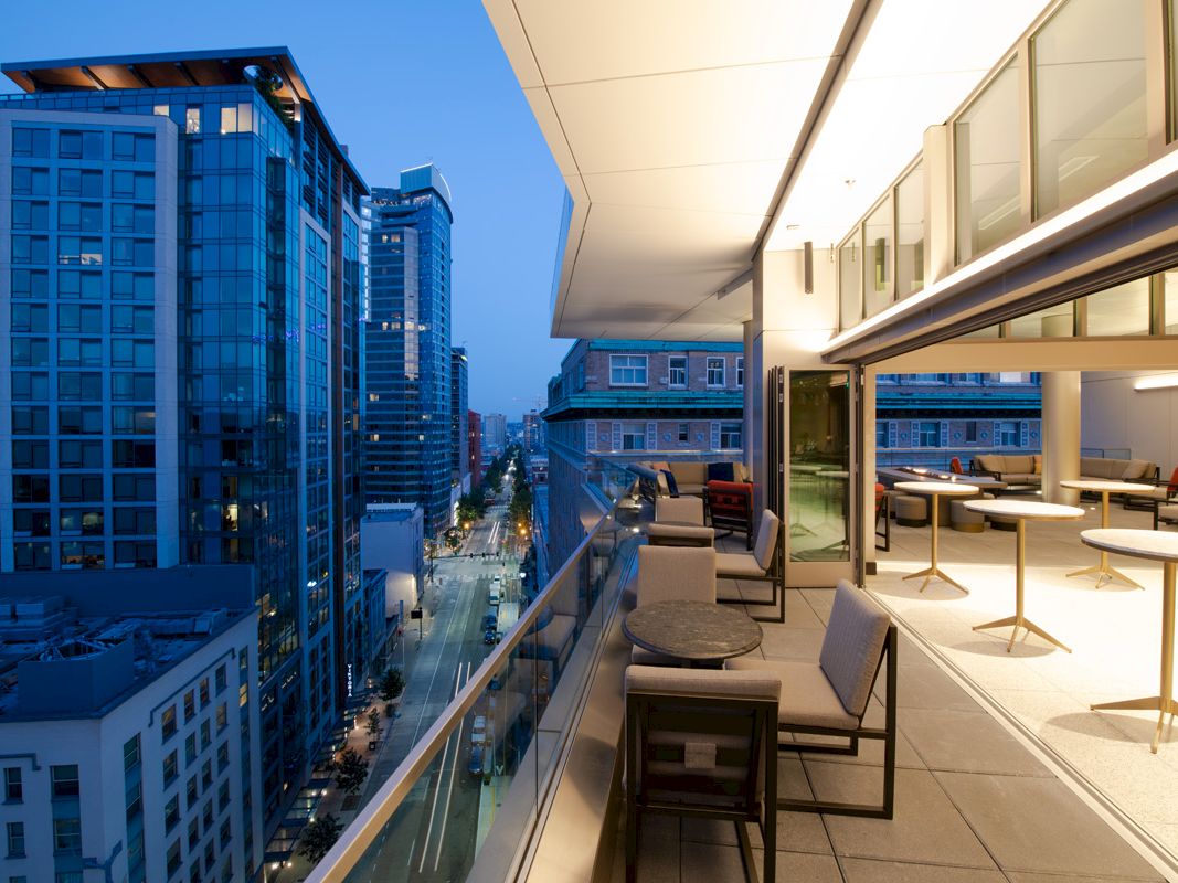 A modern city skyline viewed from a high-floor rooftop terrace with lounge seating and warm indoor lighting.